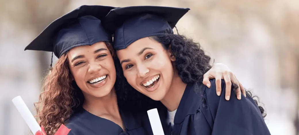 Two women embrace each other celebrating graduation in their caps and gowns while holding their degrees.