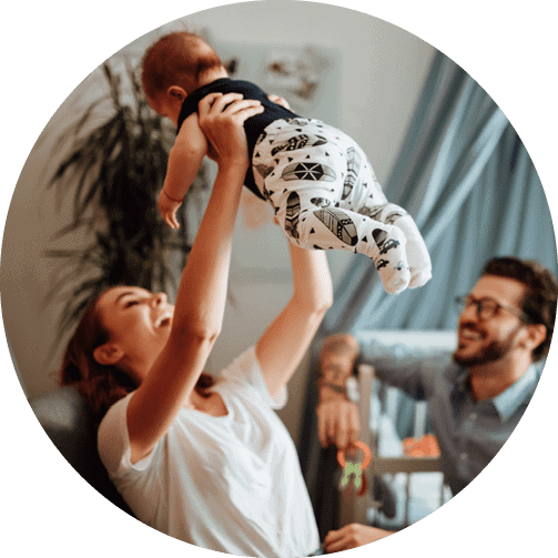 A woman in a white t-shirt lifts her baby up into the air as her husband watches, smiling from the couch.