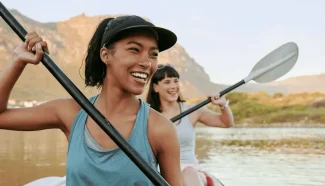 Two women are smiling while they kayak in a double-seater kayak.