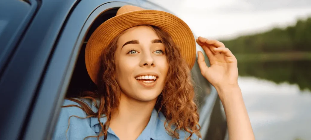 A woman with curly hair wearing a straw fedora looks out from her car window in awe of the scenery.