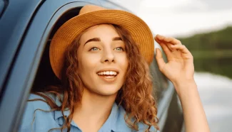 A woman with curly hair wearing a straw fedora looks out from her car window in awe of the scenery.